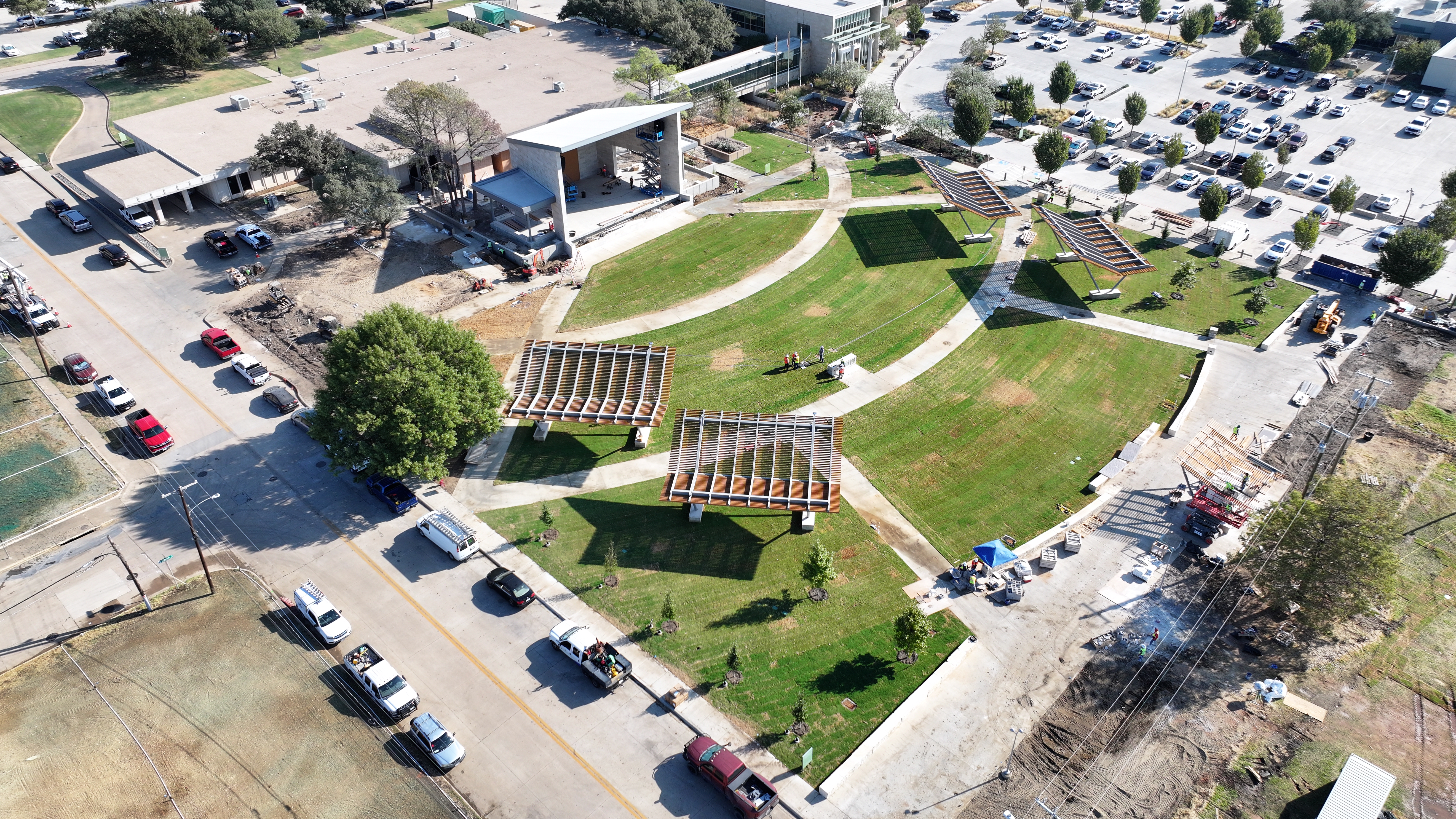 Aerial view of a green park with two wooden pergolas, winding concrete paths, and a nearby parking lot filled with cars.