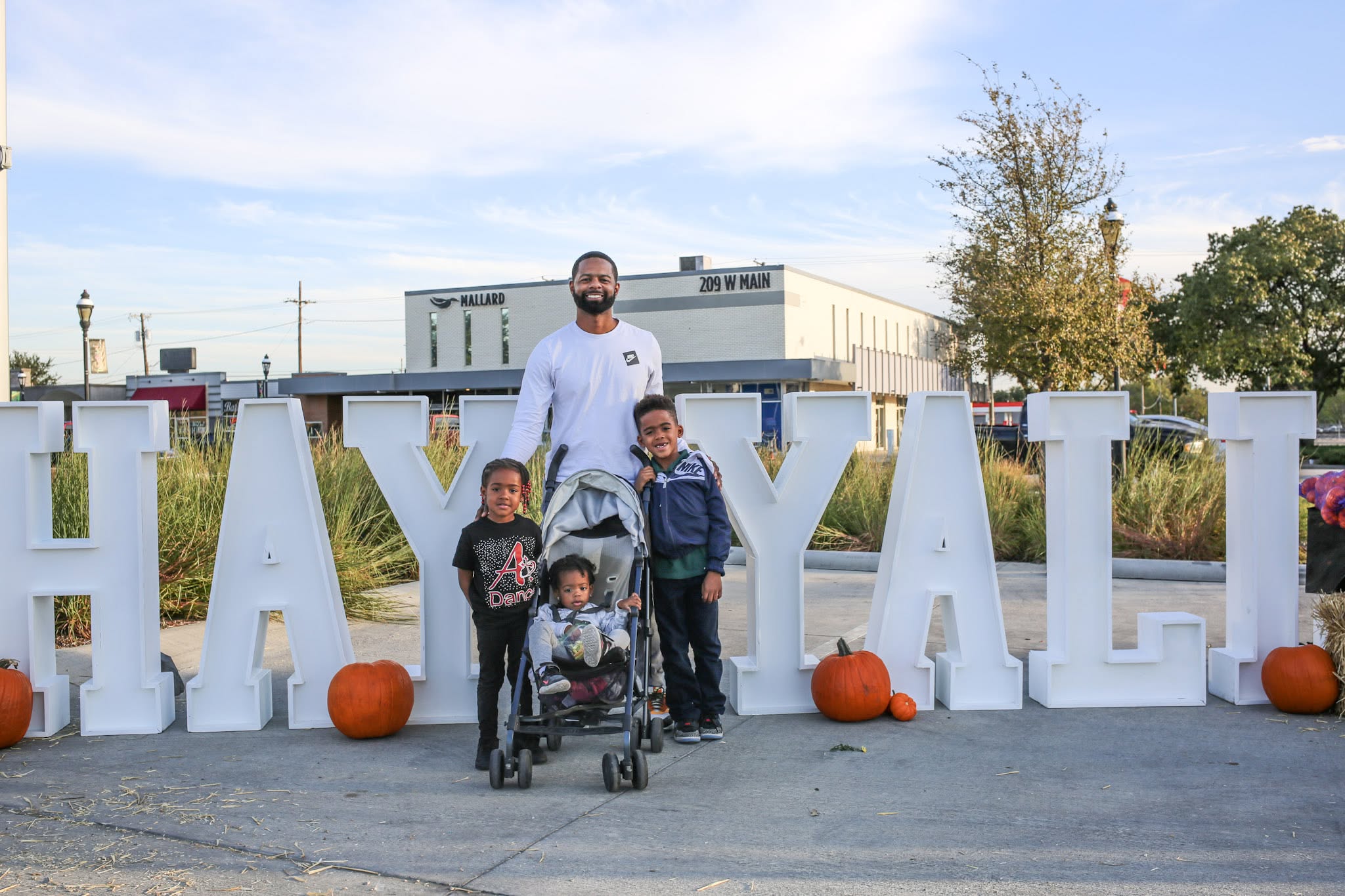 A father with three children posing in front of large white letters spelling 'HAYYALI' outdoors with pumpkins around.