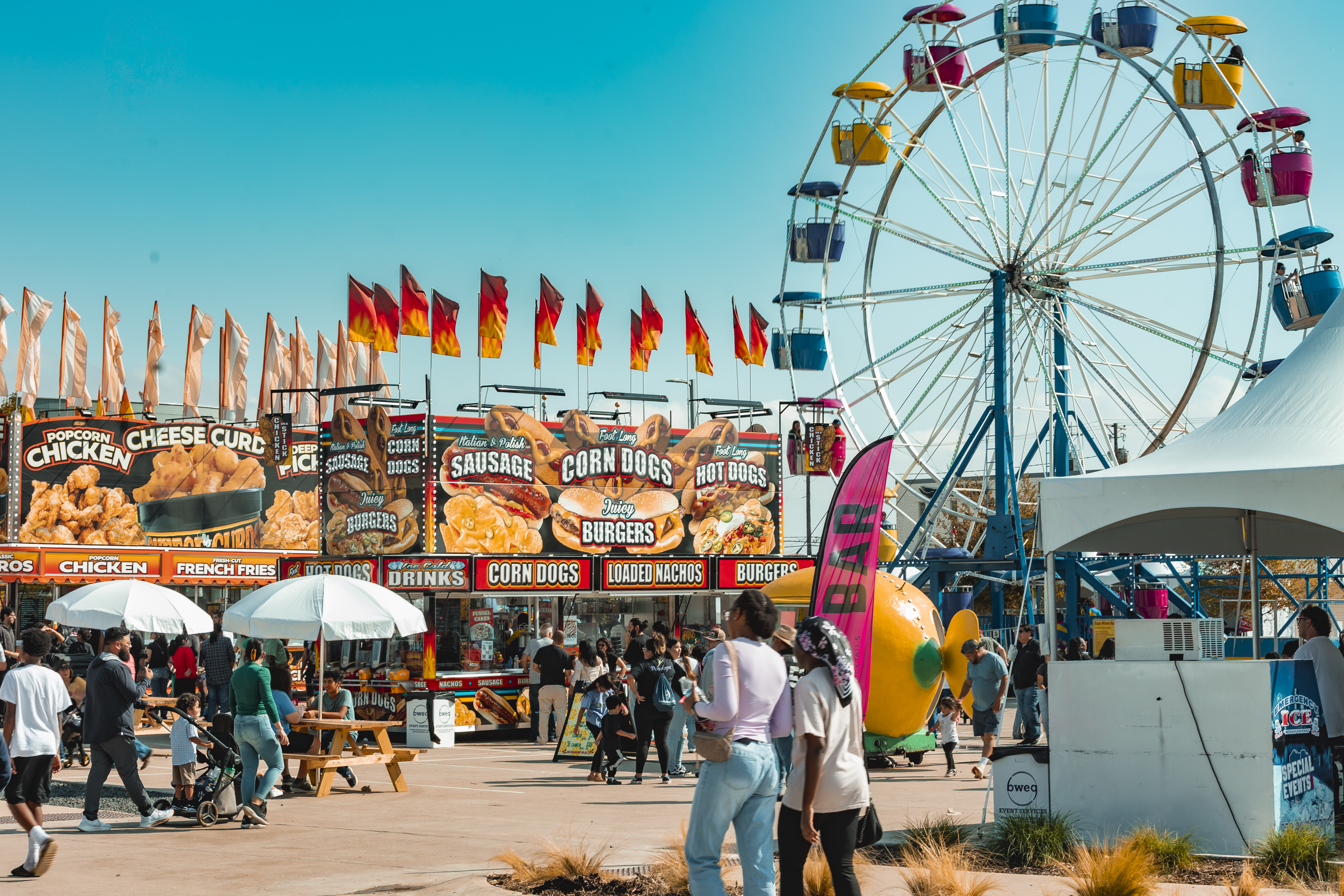 Colorful carnival food stands with flags and signs for chicken, sausage, and corn dogs under a clear blue sky.