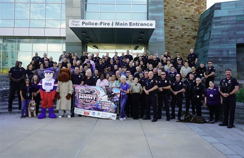 Large group of police officers and staff posing outside a building labeled Police-Fire Main Entrance with two mascots and a community event banner.