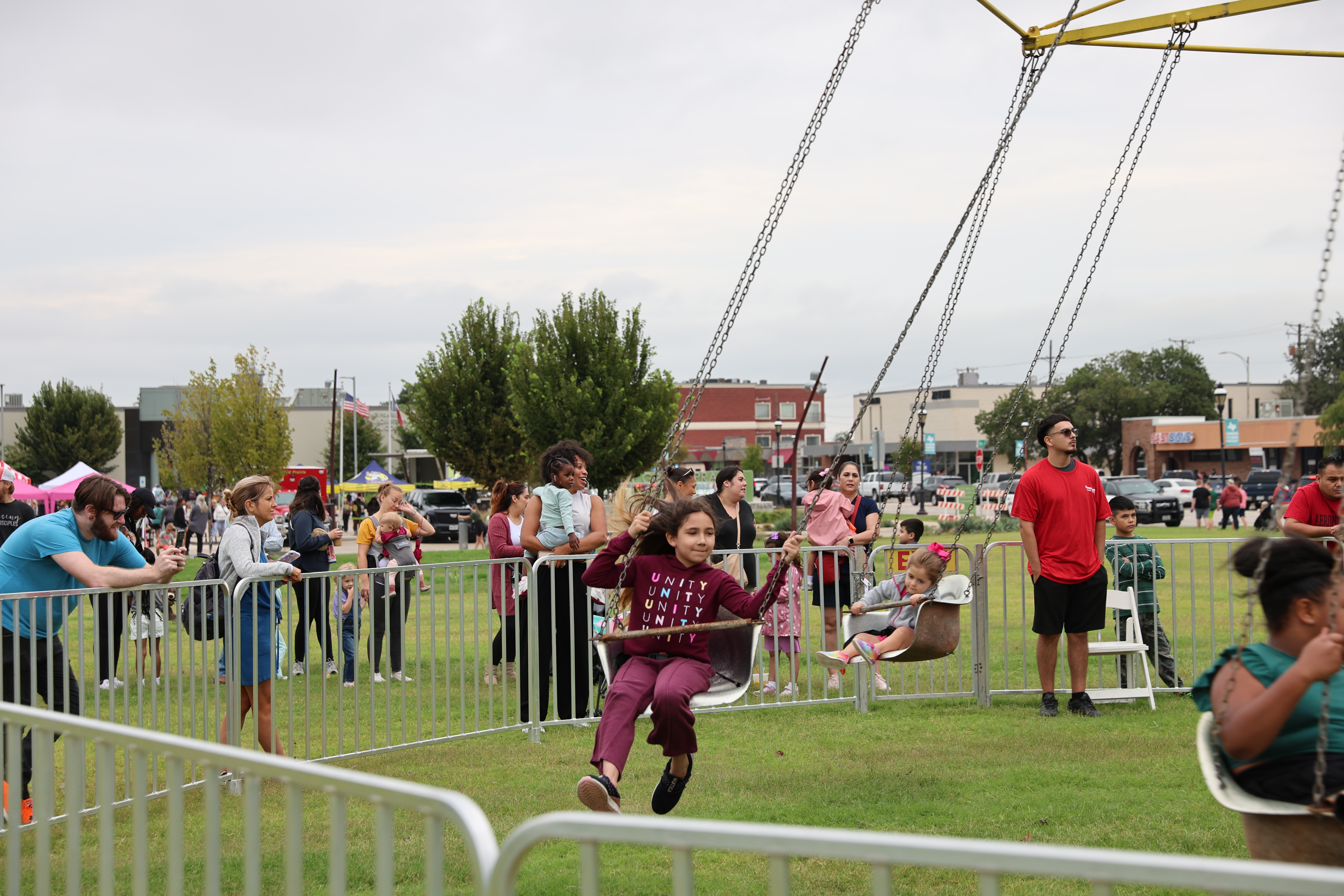 Children enjoying a swing ride at an outdoor fair with adults watching behind metal barriers on a cloudy day.