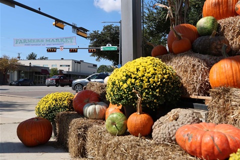 Autumn display of pumpkins, gourds, and yellow chrysanthemums arranged on hay bales at a street corner near a farmers market sign.