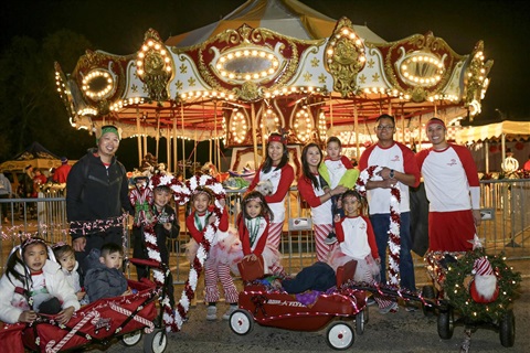 Group of children and adults dressed in festive holiday attire posing with decorated wagons in front of a brightly lit carousel at night.
