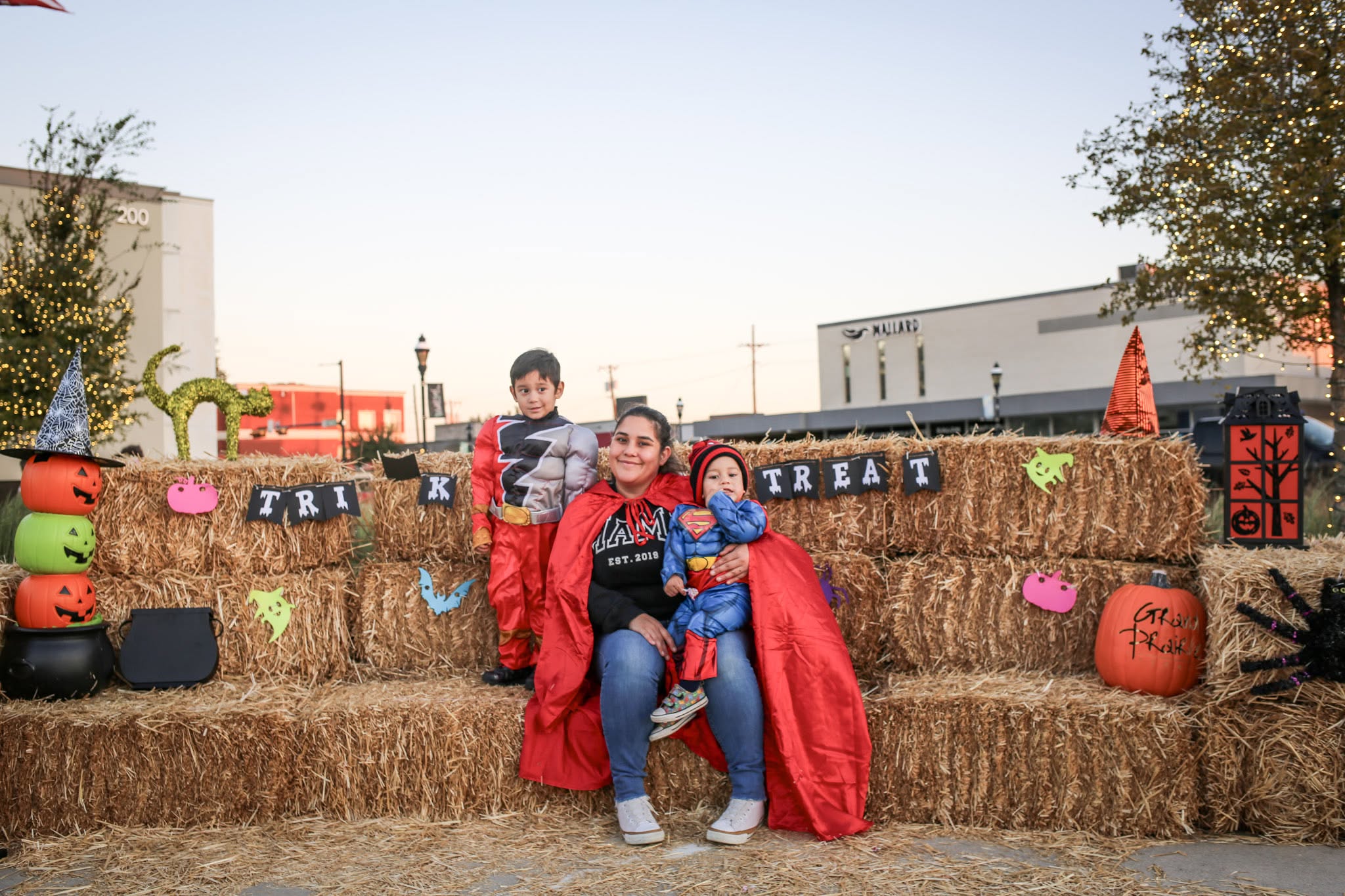 A woman and two children dressed in Halloween costumes sitting on hay bales decorated with spooky banners and pumpkins at dusk.
