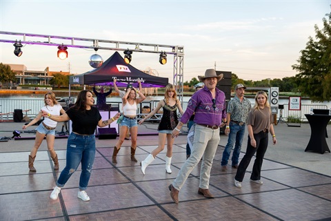 Group of people line dancing outdoors on a wooden platform near a waterfront during sunset.