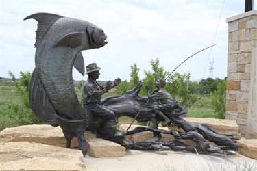 Fish Story - The Fish Story sculpture by Artist Paul Balike tells the story of a grandpa sitting on a log telling his grandson about a fish he caught. Either the grandpa or the grandson has a big imagination because the fish is 14 feet tall and 27 feet long. With the support of City Manager Tom Hart and the Grand Prairie Sports Facilities Development Corporation, the 1,000-pound sculpture helps celebrate the Lake Parks area. The Fish Story was installed at State Highway 360 and Lynn Creek Mildred Walker Parkway June 2014.