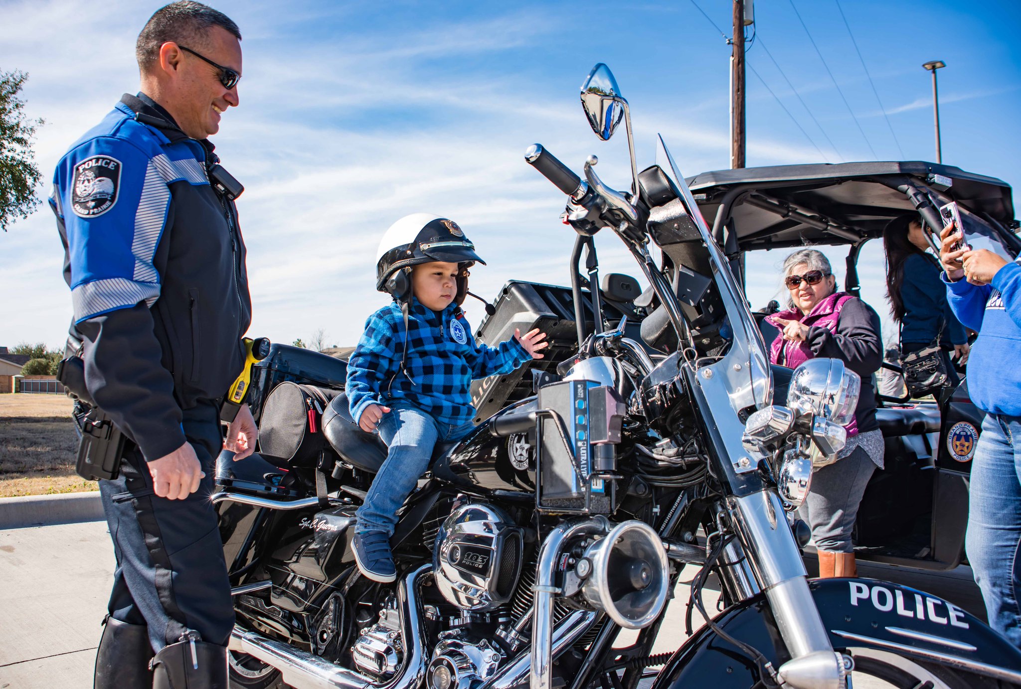 A young boy is wearing a police helmet and sitting on a police motorcycle. A police officer and the boy's parents are watching closely
