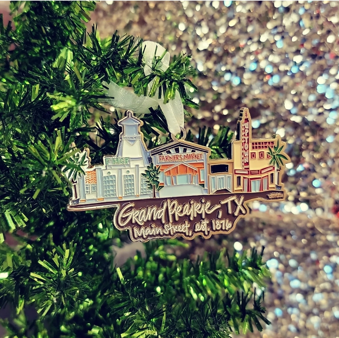 Christmas ornament hanging on a Christmas tree. The ornament is metal with a depiction of the Farmers Market and Uptown Theater on Main street. Below the picture, the ornament has the words 