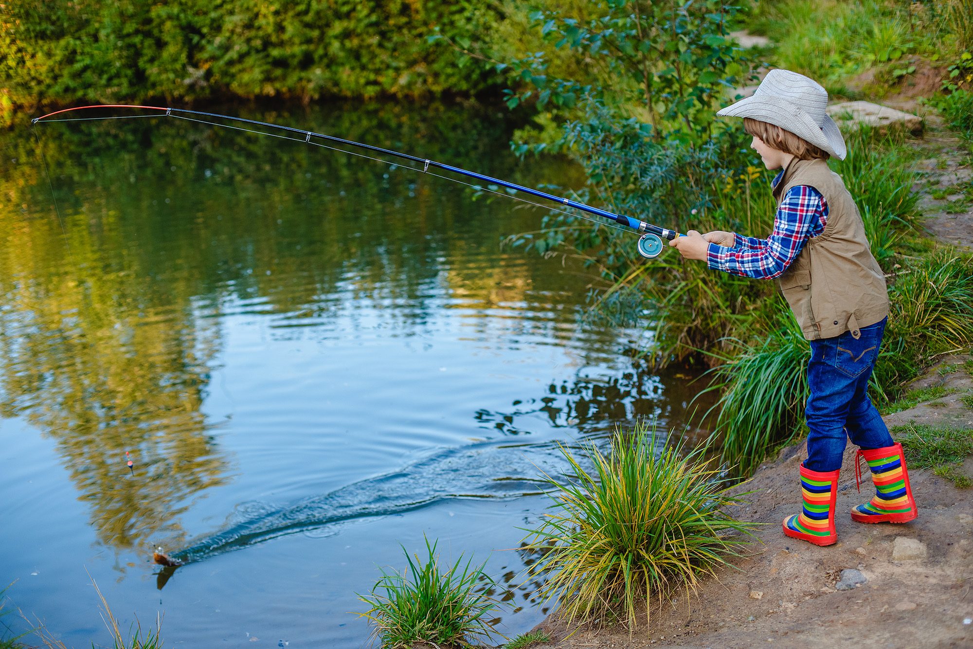 Young boy standing next to a pond. He is holding a fishing pole, and he has cast the line into the water.