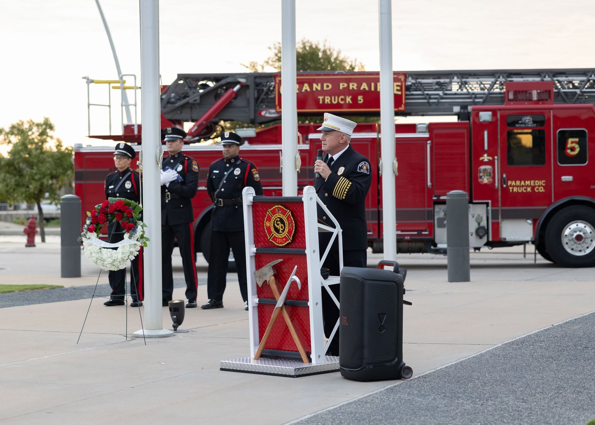 Grand Prairie Fire Chief Robert Fite at a podium decorated with a fire department emblem and crossed ceremonial axes. A memorial wreath of red and white flowers stands beside flagpoles. Behind them, several uniformed individuals stand in formation near a large red fire truck labeled “Grand Prairie Truck 5.”