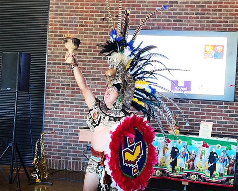 A performer in traditional Aztec ceremonial attire, holding a chalice upward while wearing an elaborate feathered headdress.