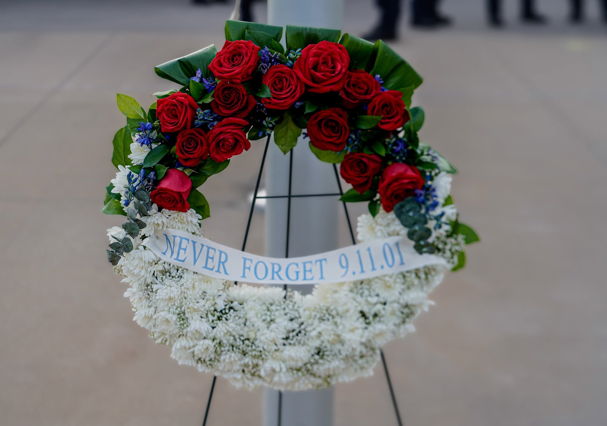 A circular memorial wreath is displayed on a stand, featuring bright red roses on the upper half and dense white flowers on the lower half, accented with green leaves and small blue blossoms. A ribbon across the center reads “NEVER FORGET 9.11.01.” The wreath is set against a smooth concrete surface.