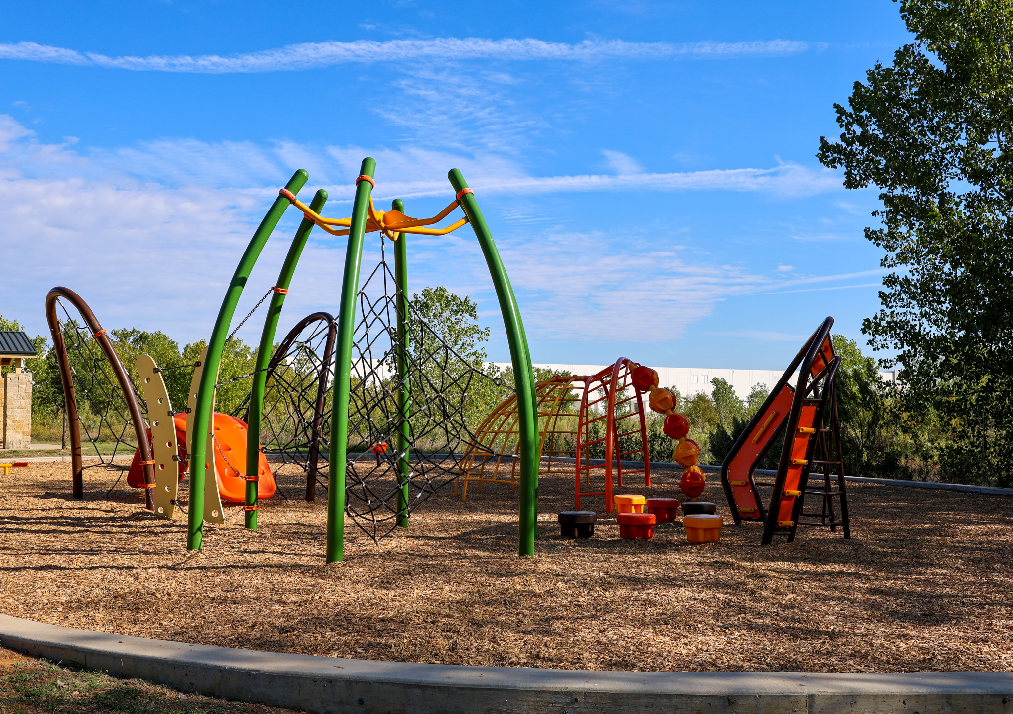 A playground in a nature park features climbing nets, slides, balance elements and tunnel structures on a wood-chip surface. Tall green support poles and colorful play equipment stand beneath a blue sky, with trees and natural vegetation surrounding the play area.