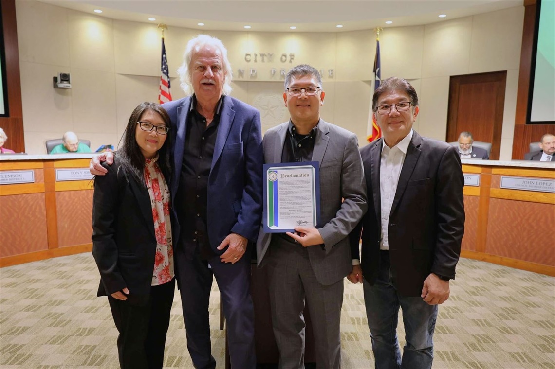 Four individuals stand together in a well-lit city council chamber. One person holds a framed proclamation. Behind them is a curved dais with seated officials, nameplates, and U.S. and Texas flags. The group stands close in the foreground, centered in front of the “City of Grand Prairie” emblem.