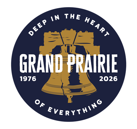 A circular navy emblem featuring a large gold Liberty‑style bell in the center. Bold white text across the bell reads “Grand Prairie.” The years “1976” and “2026” appear on either side. Curved text around the edges says “Deep in the Heart of Everything.”