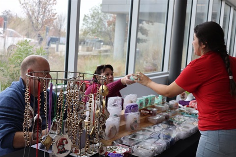 A vendor wearing a red shirt reaches across a craft display table to hand a small item to a seated woman. On the table, various handmade crafts, jewelry, and holiday ornaments hanging from a metal stand are on display next to large windows.