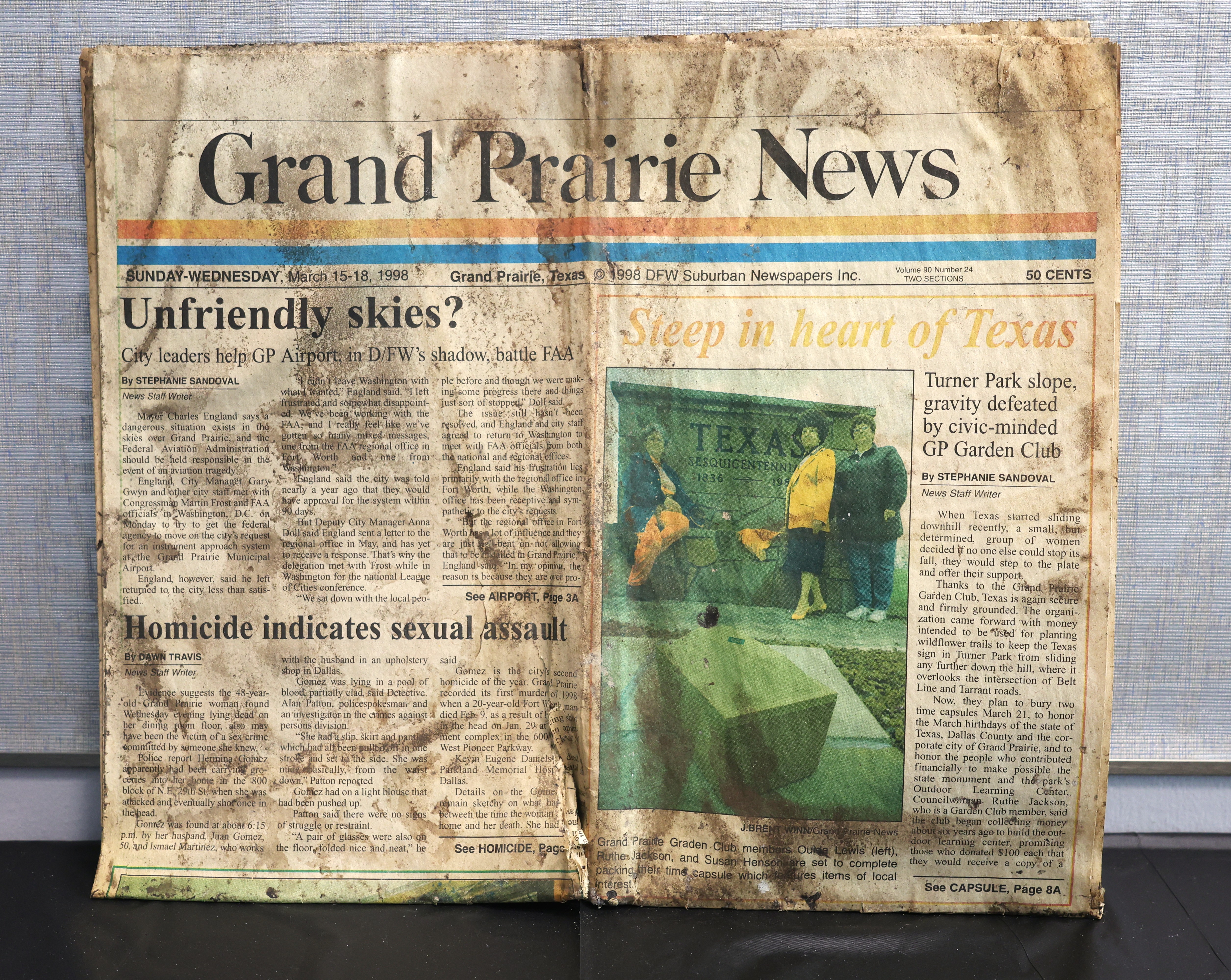 A worn, stained copy of the Grand Prairie News dated March 15–18, 1998 lies flat. The left page shows headlines about airport issues and a homicide investigation. The right page features an article titled “Sleep in heart of Texas” with a photo of three people standing beside a large Texas Sesquicentennial stone marker.  