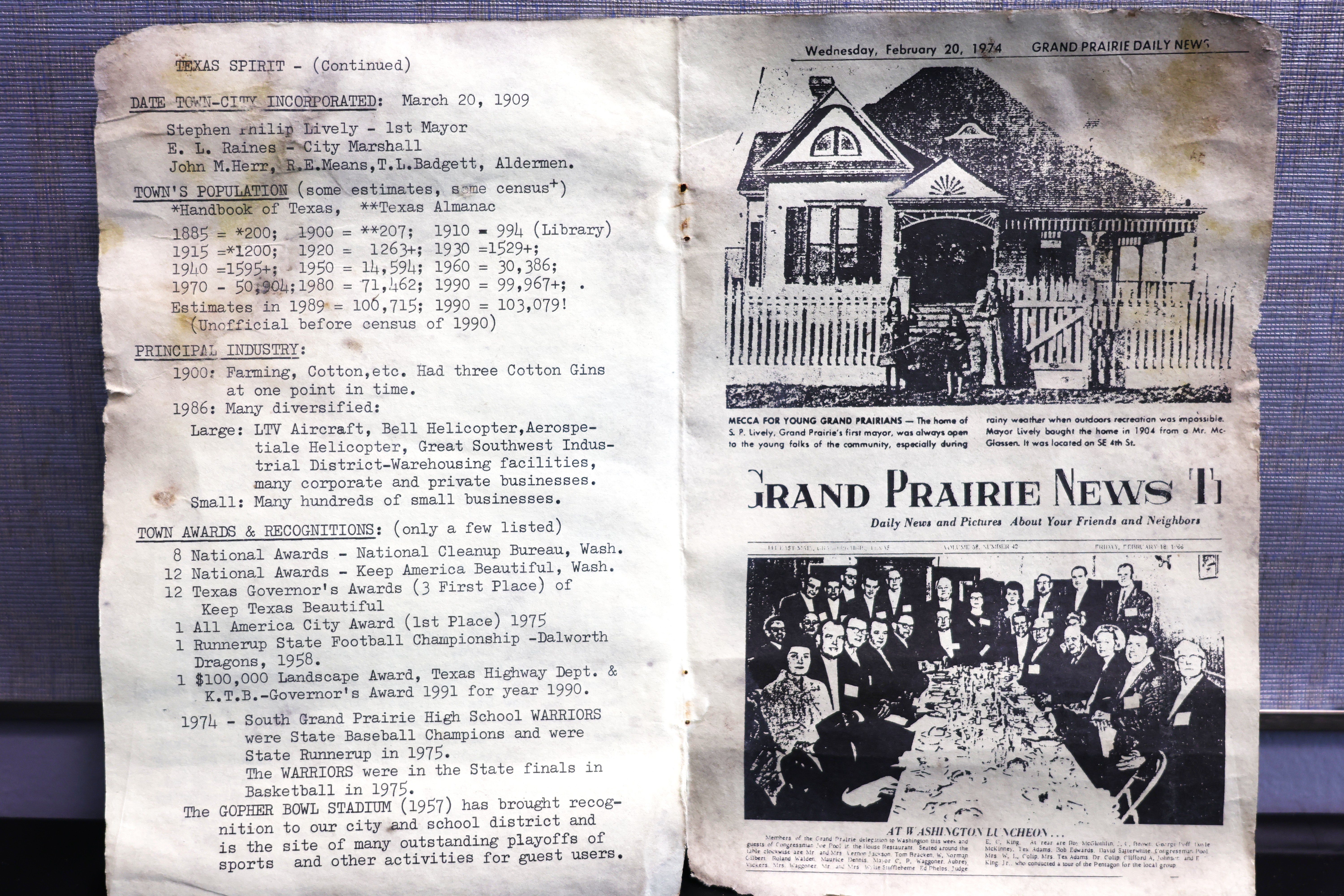 Two aged pages lie side by side. The left page lists Grand Prairie history, including incorporation in 1909, population estimates, industries, and awards. The right page is a worn Grand Prairie News front page dated February 20, 1974, showing a black‑and‑white photo of a house with a picket fence and another photo of a large formal group seated at a long table.