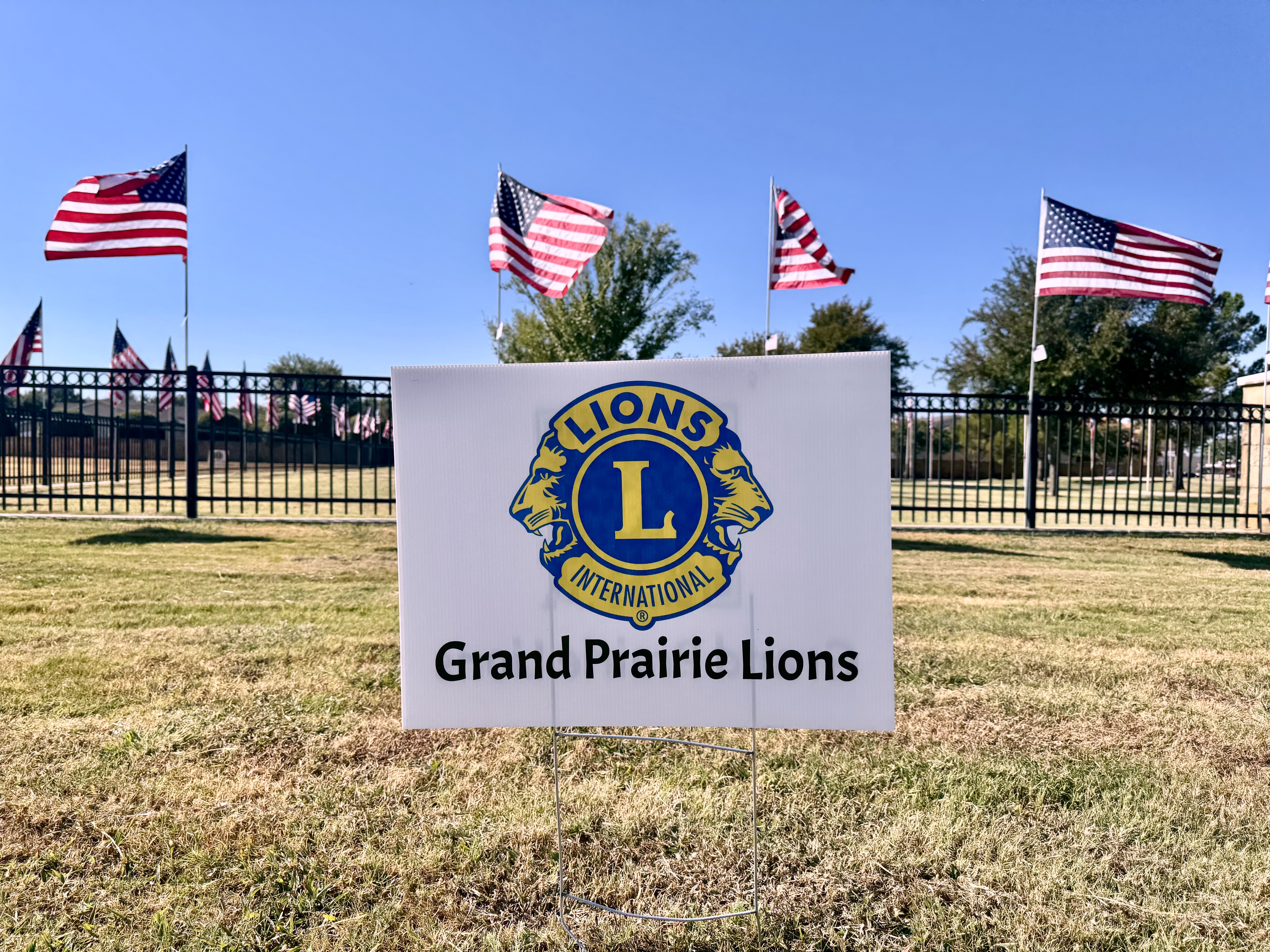 A small yard sign with the blue‑and‑yellow Lions International emblem and text reading “Grand Prairie Lions” stands on a grassy lawn. Behind it, multiple American flags on tall poles wave in the wind along a black metal fence. Trees and a clear blue sky fill the background.
