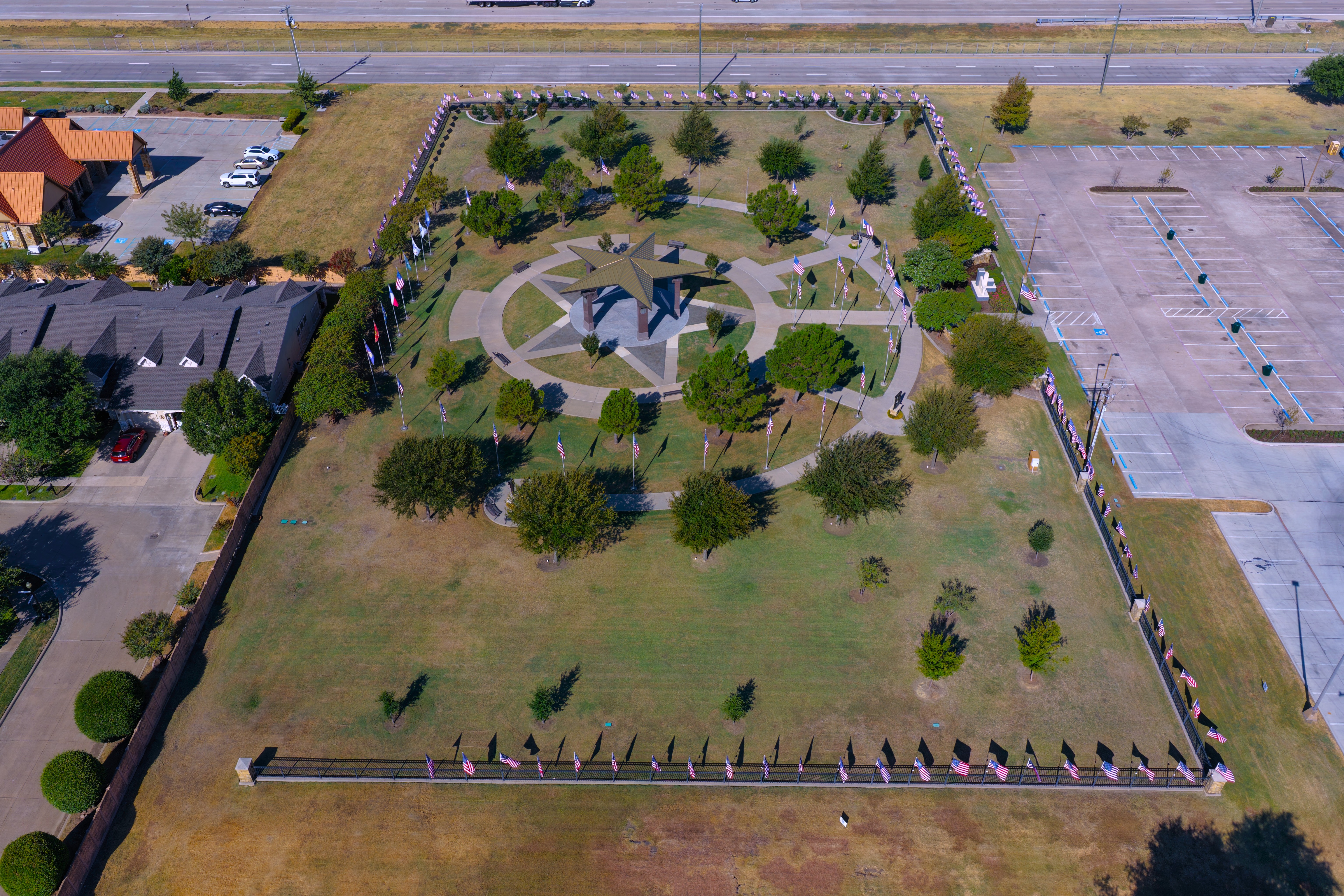 Aerial view of a large memorial park featuring a star‑shaped plaza with a central metal canopy structure. Curved walkways and evenly spaced trees surround the star. Numerous American flags line the perimeter fence and inner paths. Adjacent areas include parking lots, roads, and nearby buildings.