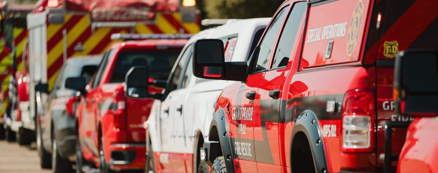 Line of emergency vehicles parked closely together, including bright red Grand Prairie Fire Department trucks and SUVs with ‘Fire Rescue’ and ‘Special Operations’ markings. A fire engine with red‑and‑yellow chevron striping is visible in the background. The vehicles are outdoors in daylight, ready for response.
