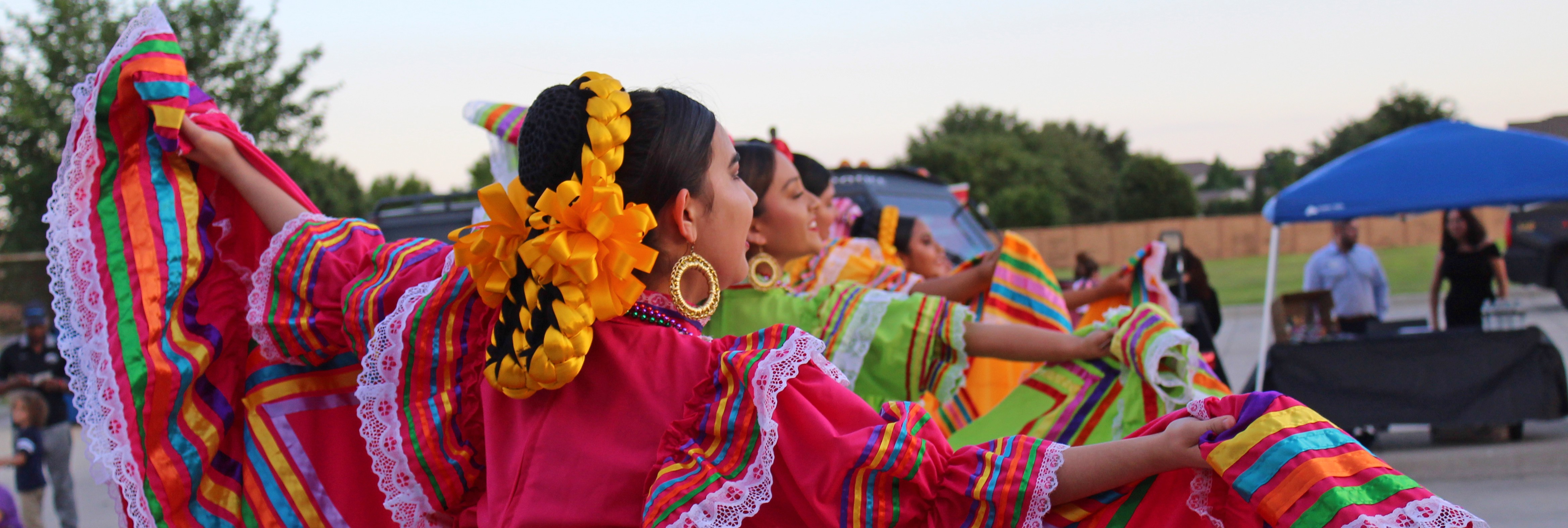 A group of dancers in brightly colored traditional Mexican dresses perform outdoors at sunset. Their wide, ruffled skirts in vivid rainbow stripes swirl as they raise their arms. Each dancer’s hair is adorned with large braided ribbons, and people watch from the background near tents and parked cars.