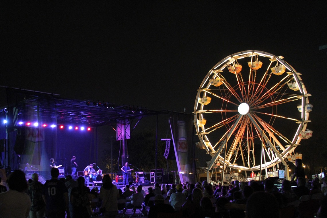 A band plays on an outdoor stage at night while a crowd watches. There is a ferris wheel near the crowd