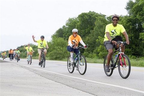 Several people, including Mayor Ron Jensen, ride their bikes down a street. People are wearing matching 