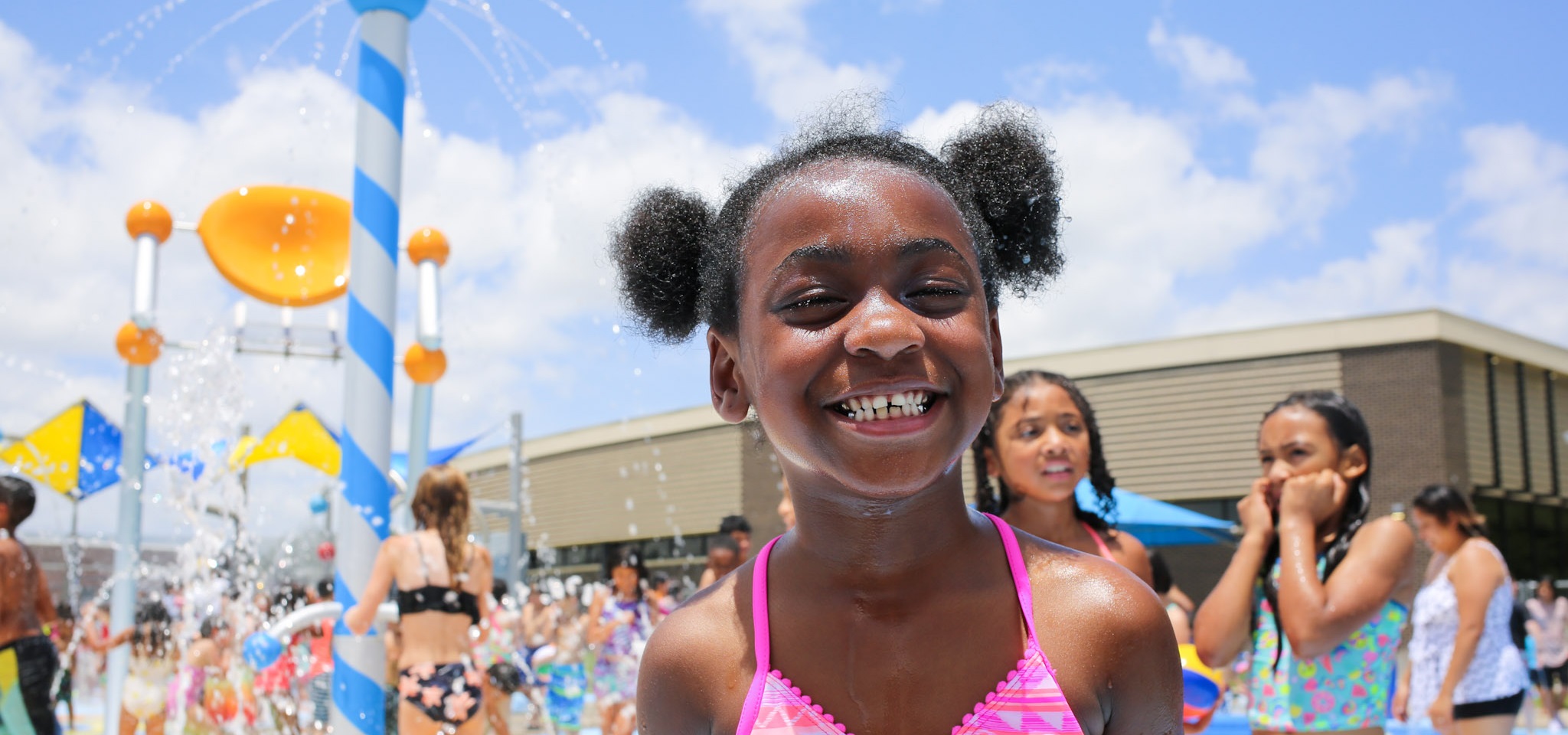 Girl standing at splash pad smiling at camera. Other kids are playing in the background.